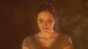 Woman with a neutral expression stands in warm, soft lighting, wearing a gray shirt.