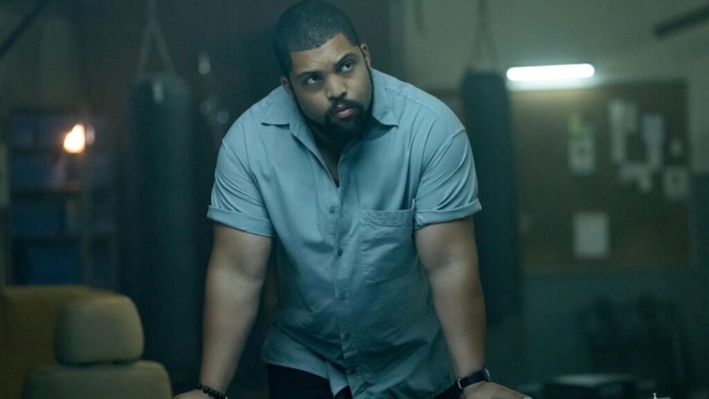A man in a light blue shirt leans on a table in a dimly lit room with blurred objects in the background.