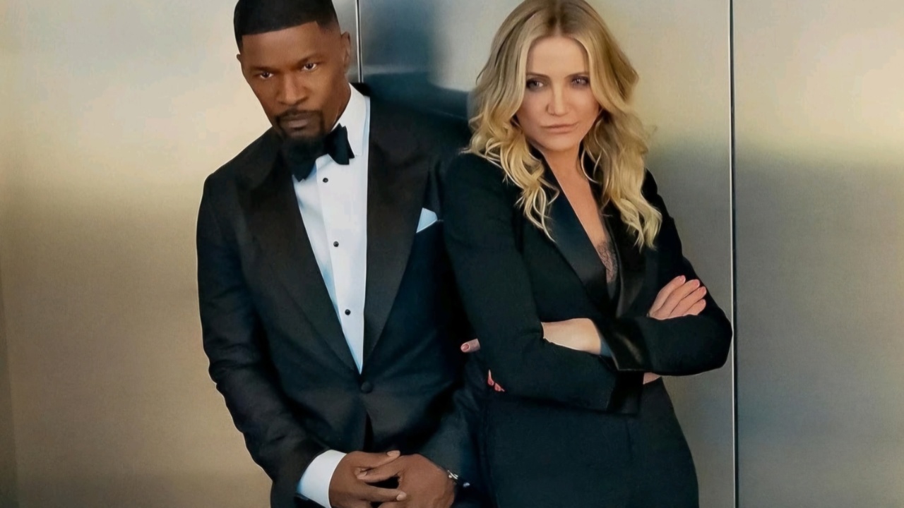 A man in a tuxedo and a woman in a black suit stand side by side against a wall.