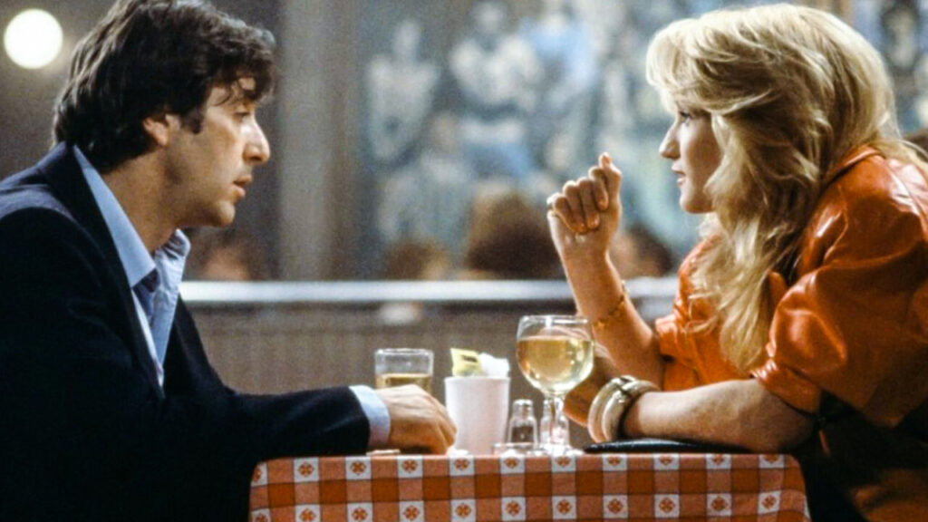 Two people sit across from each other at a checkered tablecloth-covered table, engaged in conversation. Drinks, including a glass with a lemon slice, are on the table.