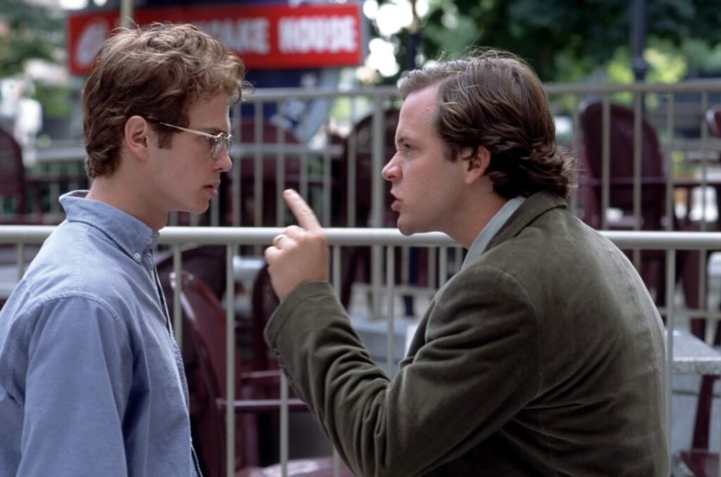 Two men stand facing each other outdoors. One man, wearing glasses, looks at the other, who is gesturing and speaking emphatically.