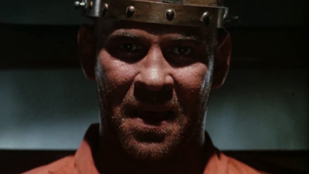 A person wearing a metal headgear appears serious, with their face partially illuminated.