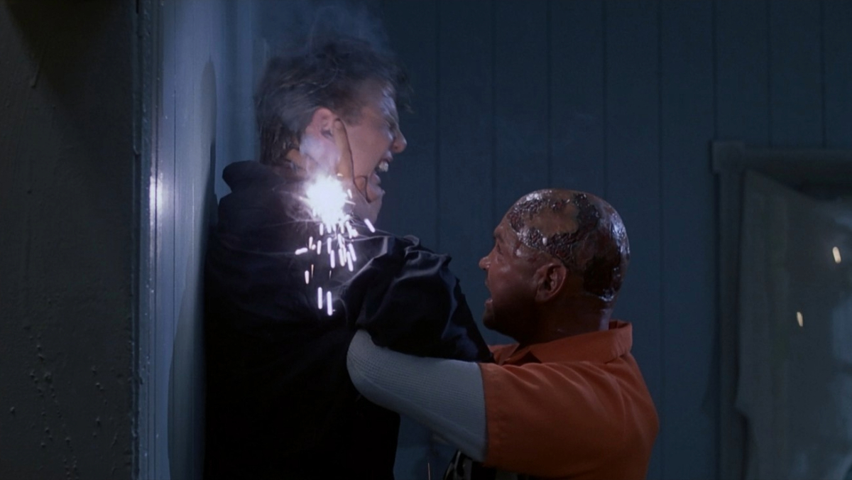 A person being pinned against a wall with visible sparks at the neck area, while another person in an orange shirt grips them.
