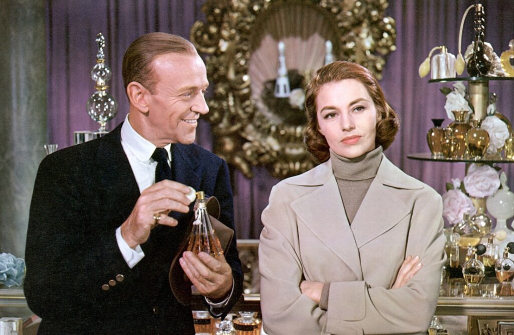 A man in a suit shows a perfume bottle to a woman with arms crossed, standing among decorative bottles and flowers, against a purple curtain backdrop.