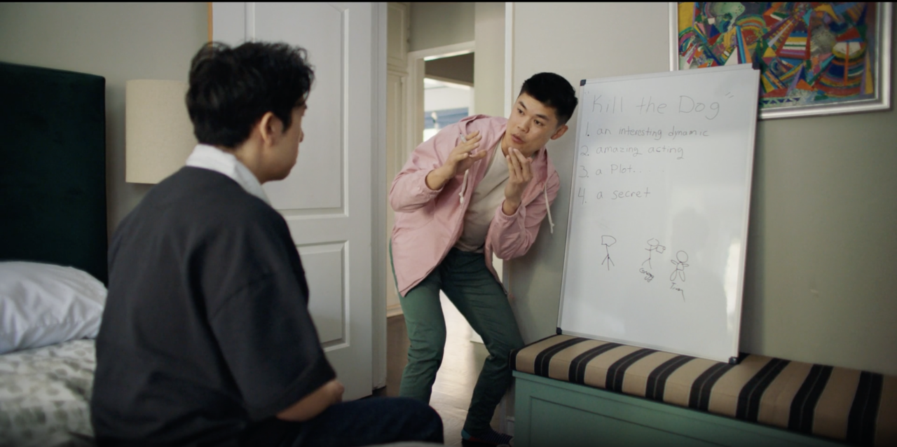 Two people in a room discuss ideas written on a whiteboard. One person stands gesturing near the board, while the other sits on a bed observing.