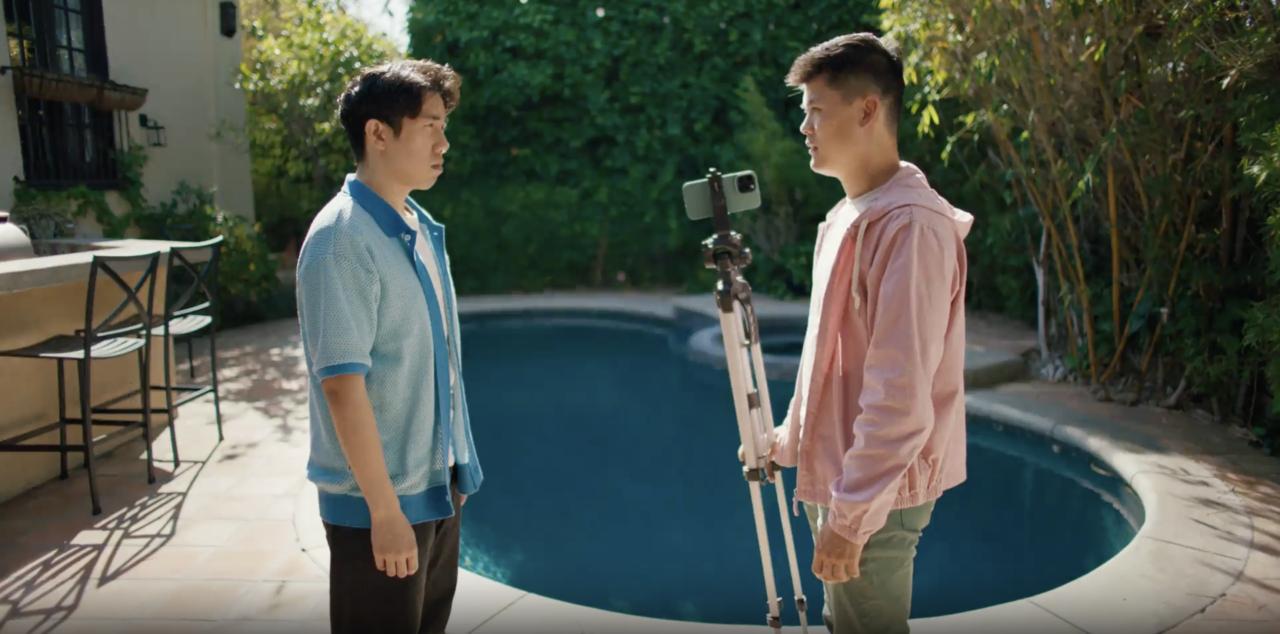 Two people stand facing each other by a pool. One holds a camera on a tripod. Outdoor setting with greenery and patio furniture.