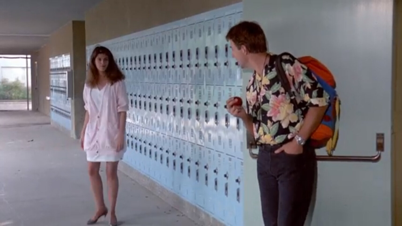 A person in a pink outfit and another with a floral shirt and backpack stand in a school hallway near lockers. One is holding an apple.