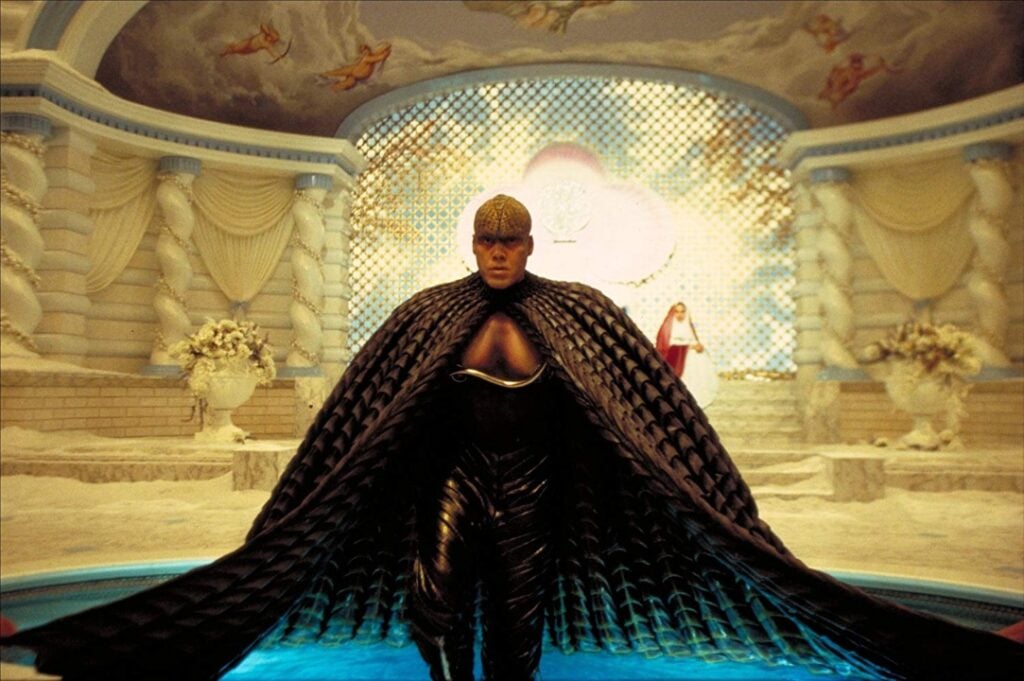 A person in an ornate costume with a scale-like cape stands in a luxurious, decorated room with a mosaic wall and drapery.