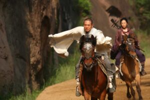 Two people riding horses on a dirt path; the leading rider wears a white flowing garment, while the second rider carries a musical instrument on their back.