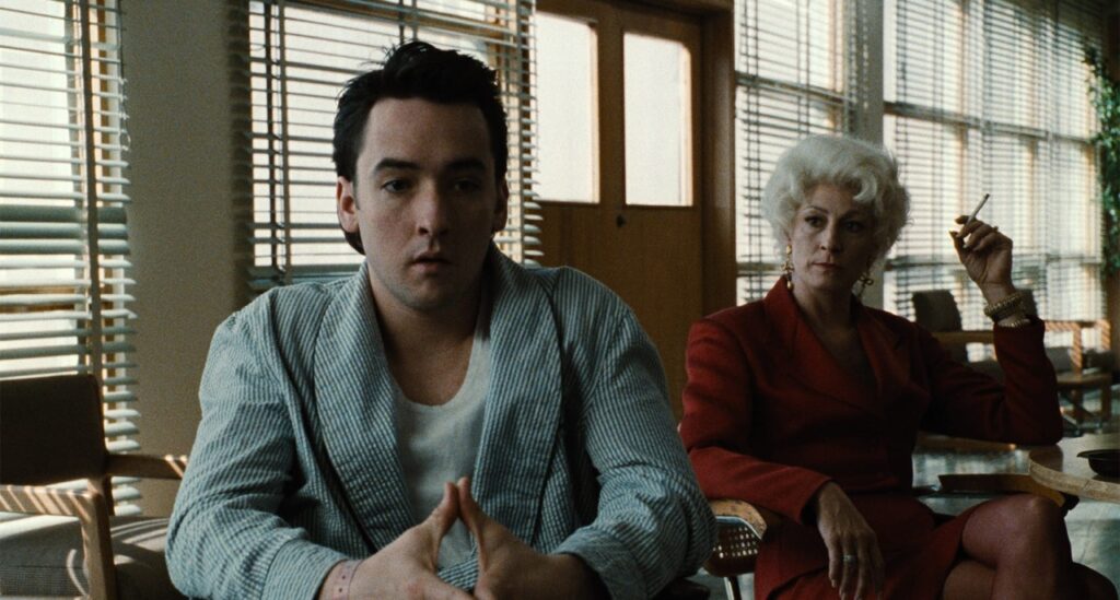 A man in a striped robe sits pensively beside a woman in a red suit in a sunlit room with blinds.