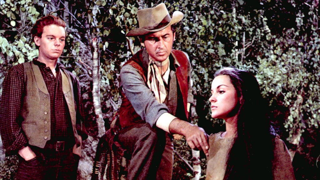 Three people in period clothing stand in a wooded area. One man kneels with his hand on a woman's shoulder, while another man looks on.