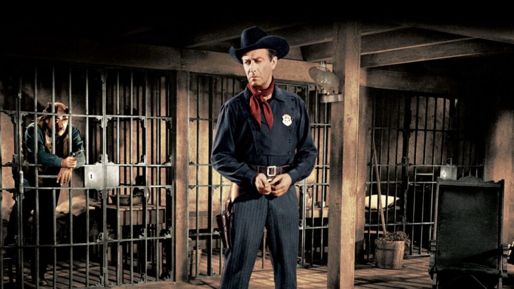 A man wearing a sheriff's badge stands in a Western-style jailhouse, while another person is seated behind bars.