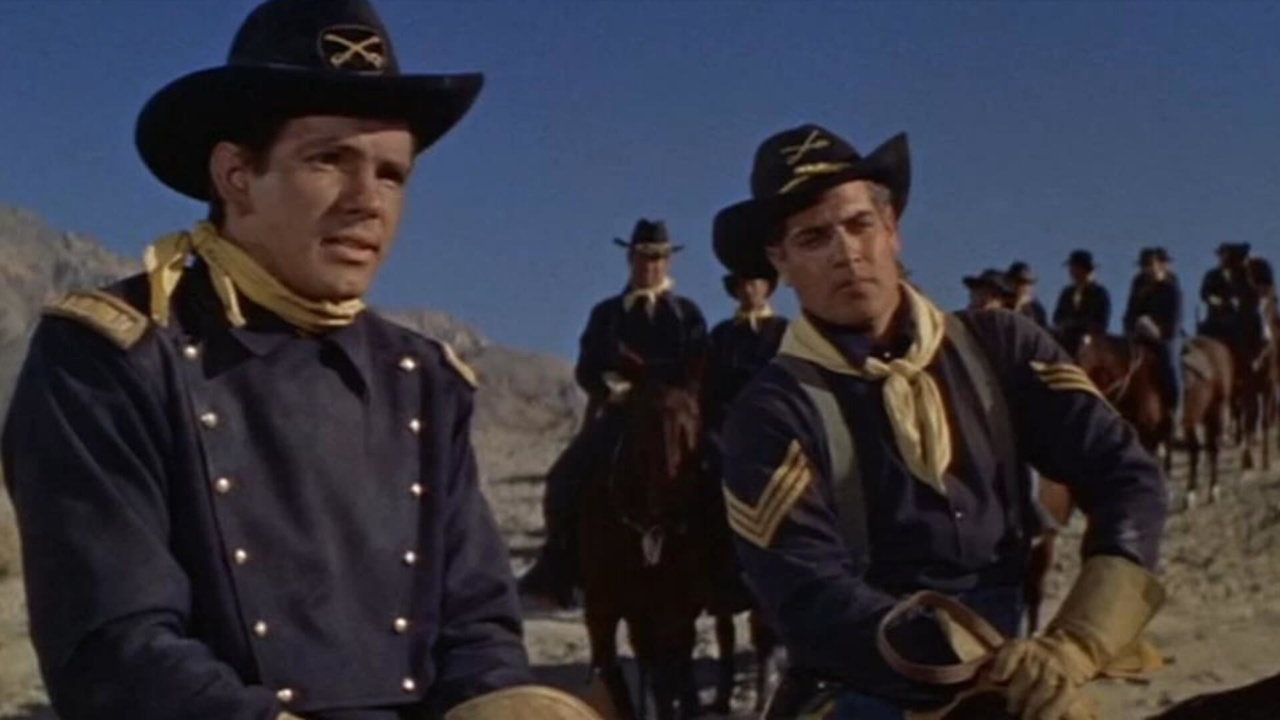Two men wearing cavalry uniforms and hats ride horses, with more riders in similar attire visible in the background, set against a clear blue sky.