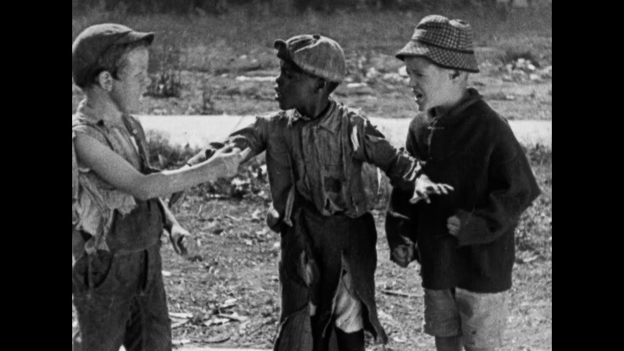 Three children standing together outdoors; one in tattered clothes gesturing to another, while the third child watches. The scene is black and white.