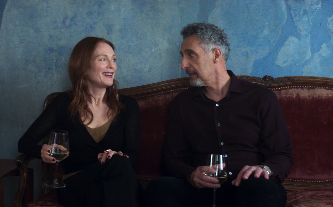 Two people sitting on a couch, both holding a glass of wine, engaged in conversation against a blue-toned wall backdrop.