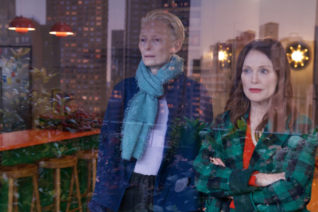 Two women with serious expressions stand indoors, looking out a window. The background shows reflections of an urban cityscape and indoor plants.
