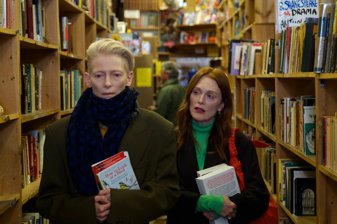 Two people are standing in a bookstore aisle, holding books. Shelves are filled with books around them, and other people are visible in the background.
