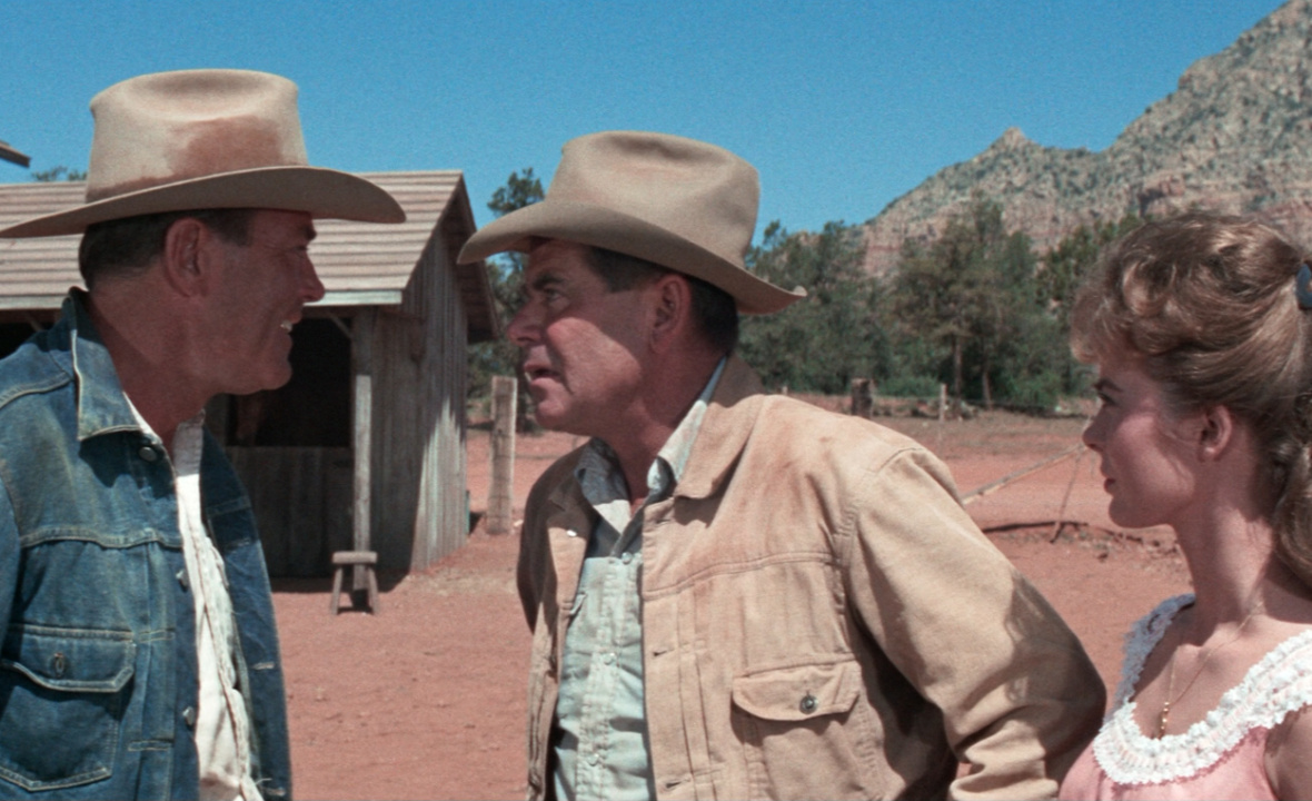 Three people stand outdoors; two men in cowboy hats face each other, while a woman looks on. A wooden building and desert landscape are in the background.