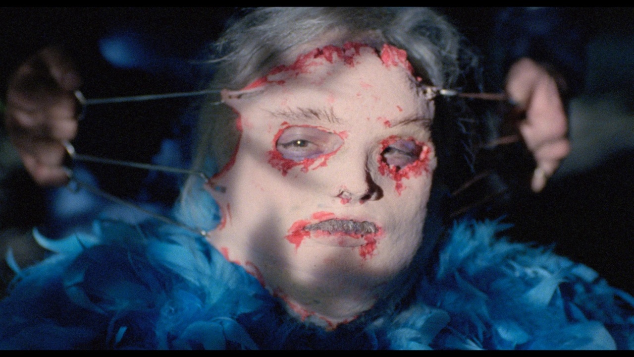 A person with a mask partially attached, showing red and raw edges, stares forward amidst a blue feathered collar in dim lighting.