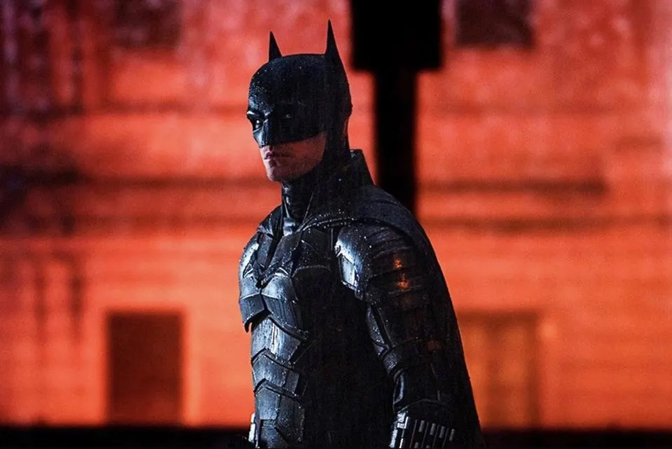 A person in a black armored suit with bat-like ears stands in front of a blurred orange background.