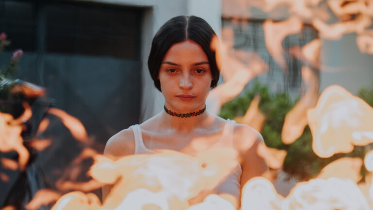 A person with a choker stands behind flames, looking forward with a neutral expression. The background is blurred.