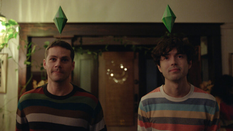Two people wearing striped sweaters stand indoors, each with a green diamond-shaped object above their head, resembling icons from The Sims video game.