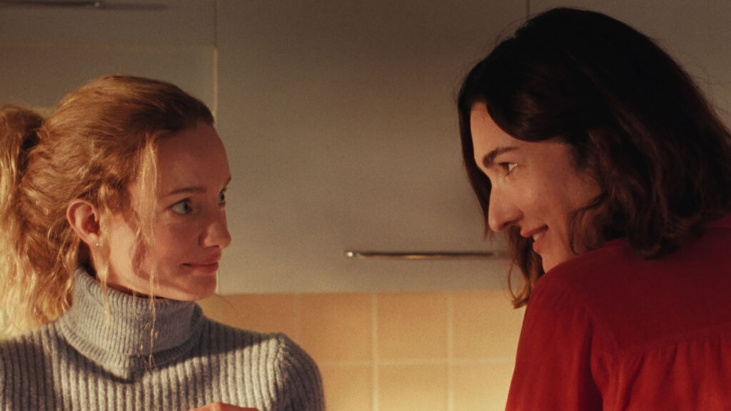 Two individuals are standing in a kitchen, facing each other. The person on the left wears a gray turtleneck, and the person on the right wears a red shirt. They appear to be engaged in conversation.
