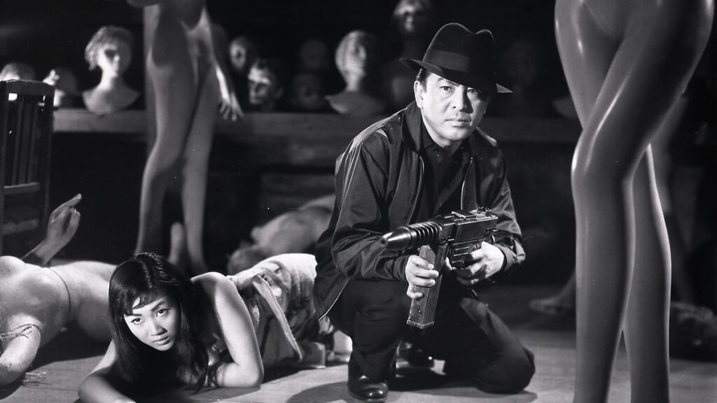 A man in a fedora holds a gun while crouching beside a woman on the floor, surrounded by mannequin parts in a dimly lit room.