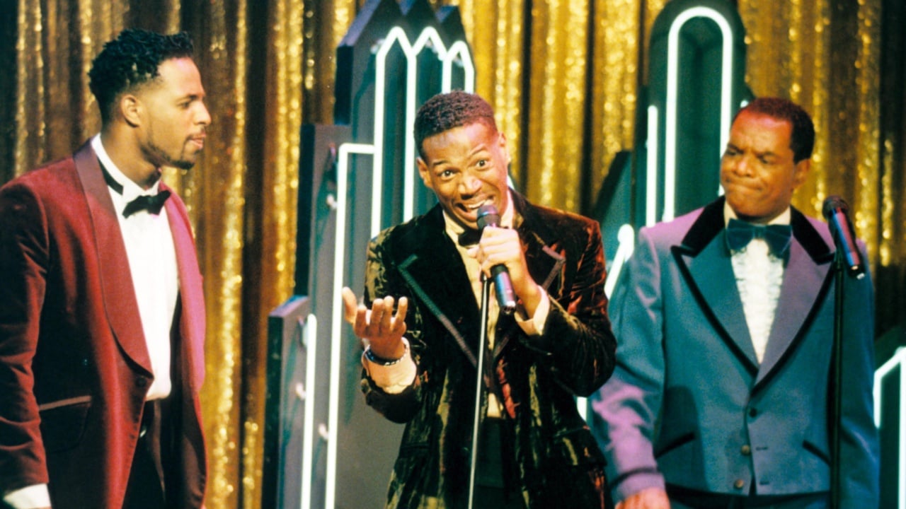 Three men in formal attire stand on stage against a backdrop of gold curtains and decorative arches. The man in the center speaks into a microphone, while the others look on.