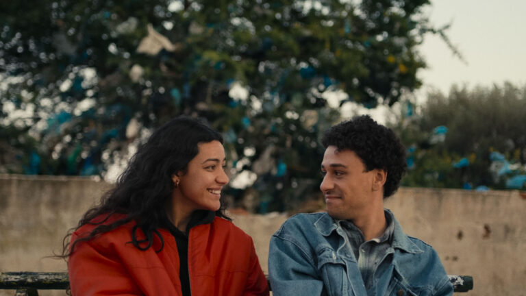 Two people sitting on a bench outdoors, smiling at each other. One wears a red jacket, the other a denim jacket. Trees and a wall are in the background.
