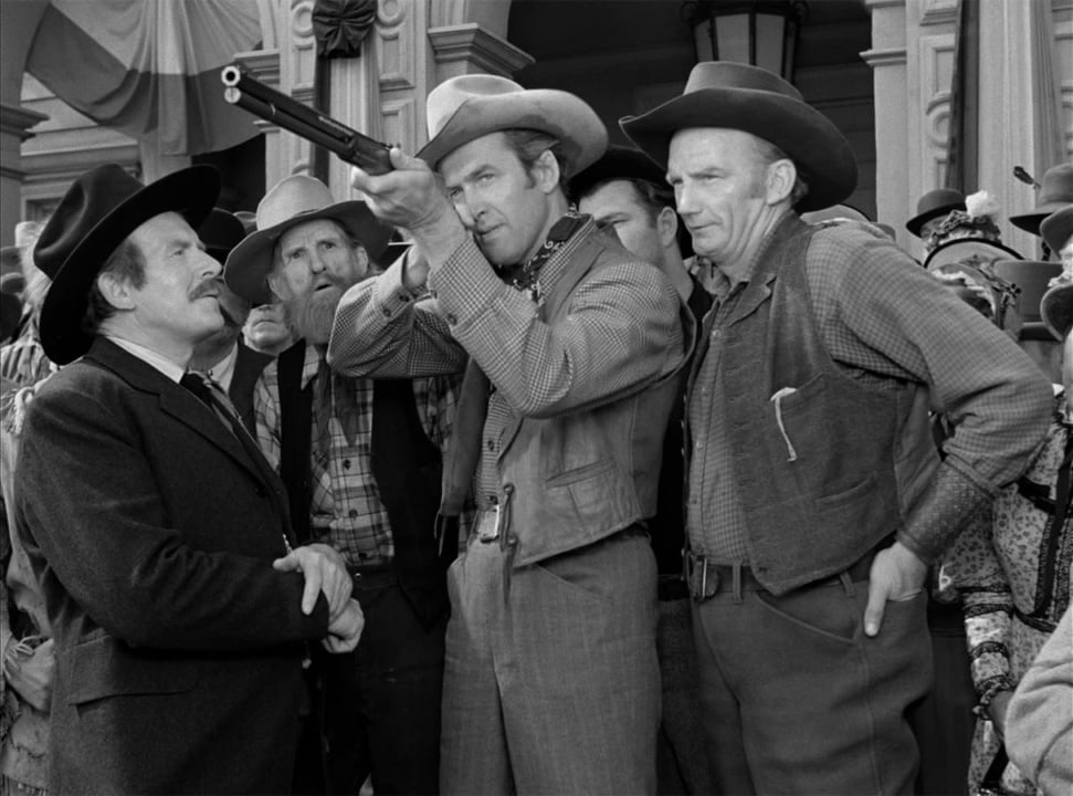 A group of men in Western attire gather outdoors. One man points a shotgun upwards while others watch.