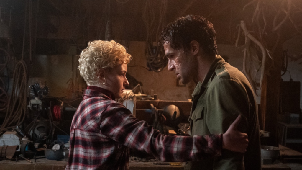 Two people stand in a cluttered workshop, facing each other closely. The person on the left wears a plaid shirt, the person on the right wears a green jacket. Both appear serious.