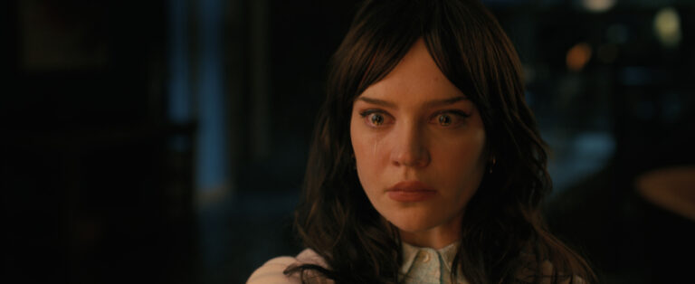 A woman with dark hair is looking forward with a serious expression, eyes slightly teary, in a dimly lit room.
