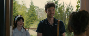 A man and woman stand at a doorway, smiling. Trees and bushes are visible in the background.