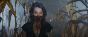 A person stands in a cornfield with blood on their face, wearing a dark shirt, against a foggy background.