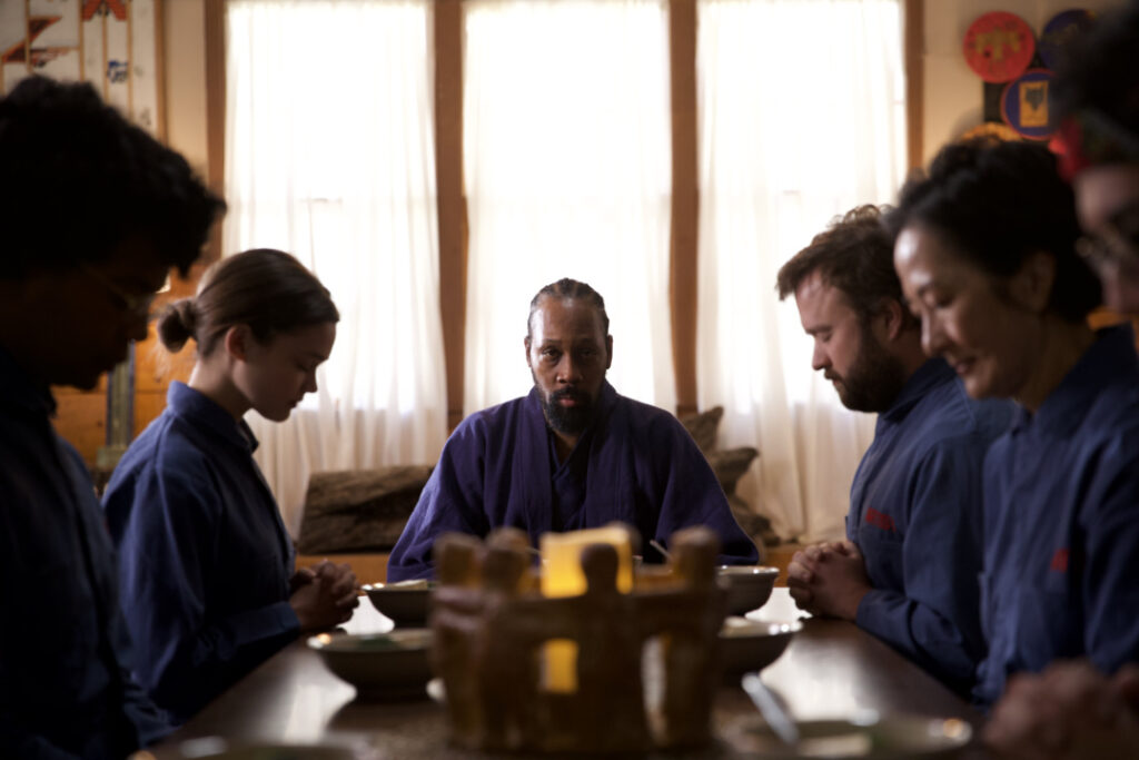 A group of people in blue robes sit at a dining table, heads bowed in contemplation or prayer, with a lit candle in the center.