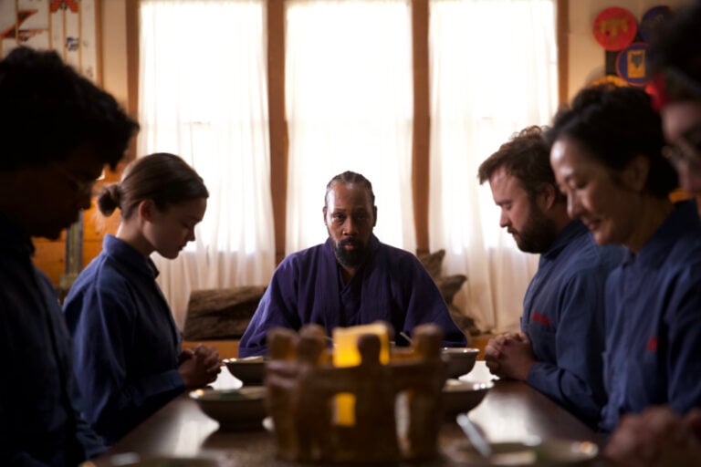 A group of people in blue robes sit at a dining table, heads bowed in contemplation or prayer, with a lit candle in the center.