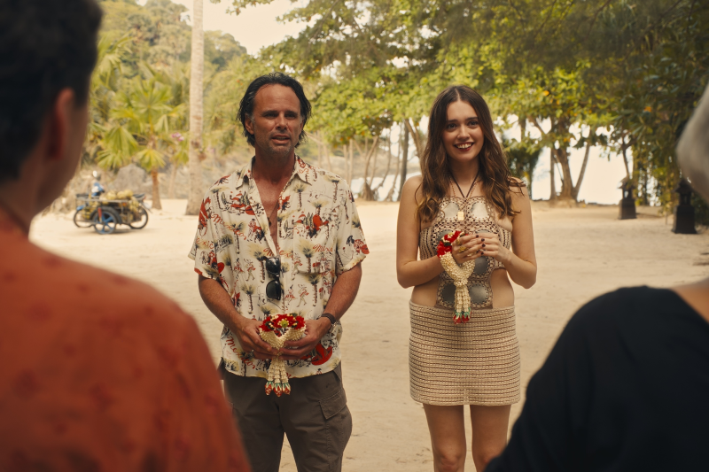 Two people stand outdoors holding festive chicken sculptures with flowers. The man wears a patterned shirt; the woman wears a crochet outfit. Trees and a motorbike are in the background.