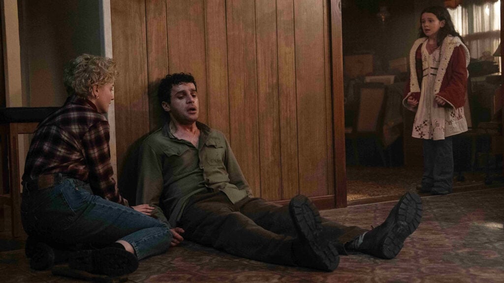 A woman kneels beside a seated man on the floor, while a girl with a concerned expression stands nearby in a dimly lit room.