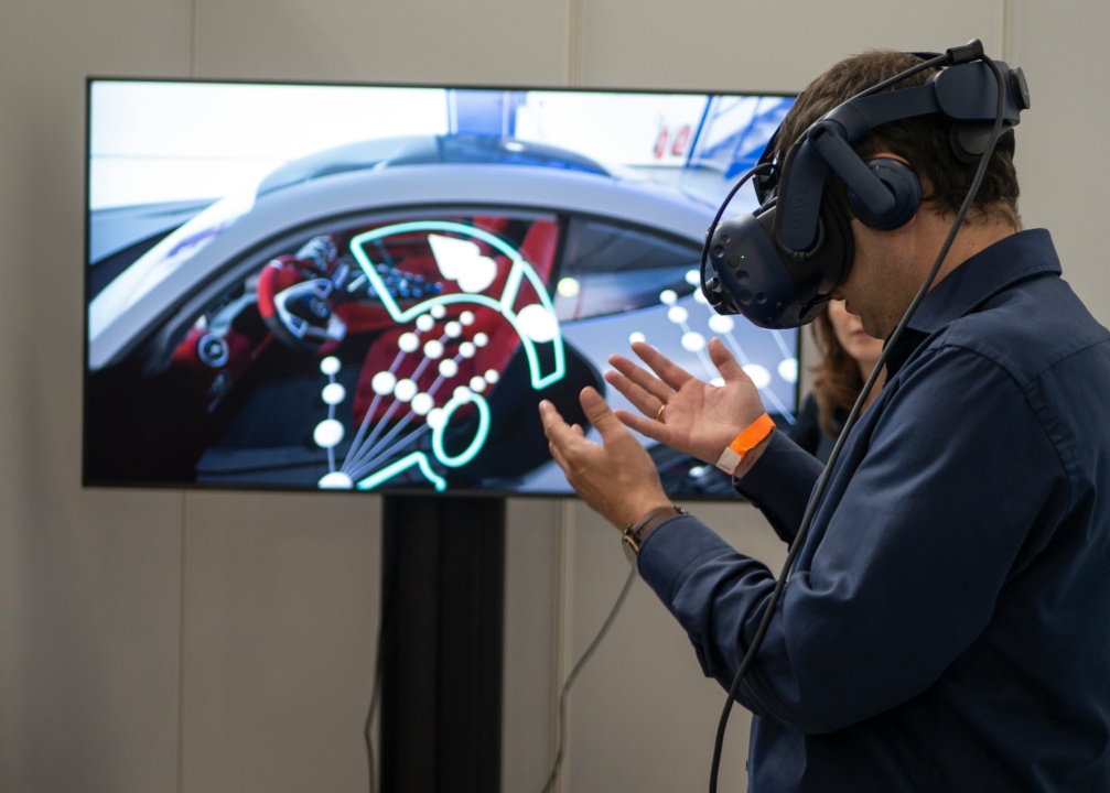 A person wearing a VR headset interacts with a virtual car dashboard displayed on a screen.