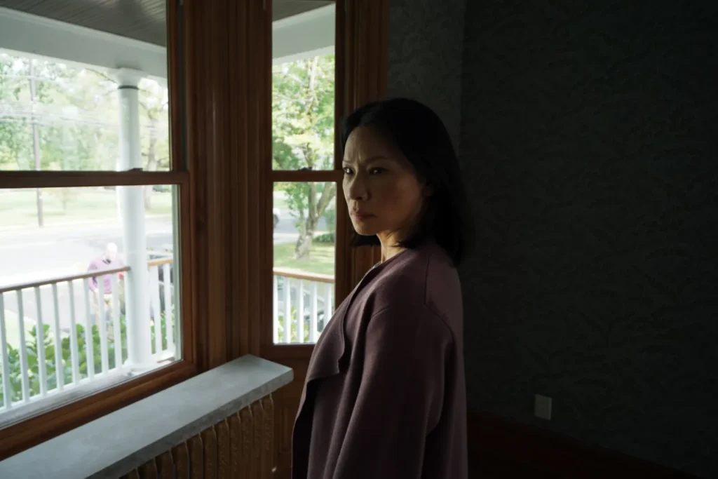 A woman in a purple coat stands by a window, looking outside. The room is dimly lit, and a porch with white railings is visible through the window.