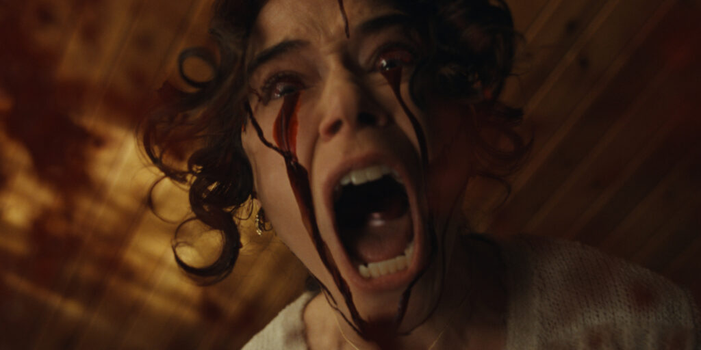 A person with curly hair is screaming, with streams of dark liquid running from their eyes against a blurry wooden background.