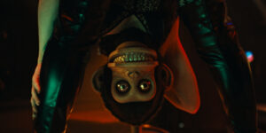 A person in black attire is upside down, holding a toy monkey with big eyes and a wide grin, illuminated by red and green lights.