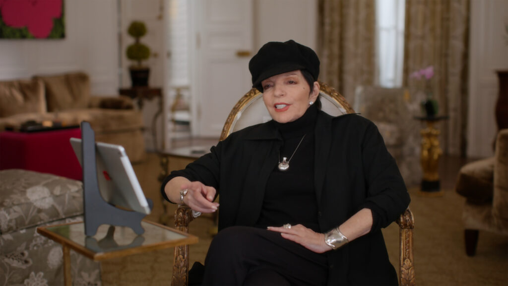 Person in black outfit and hat sits in an ornate chair, speaking and gesturing with a tablet on a stand nearby in a decorated room.