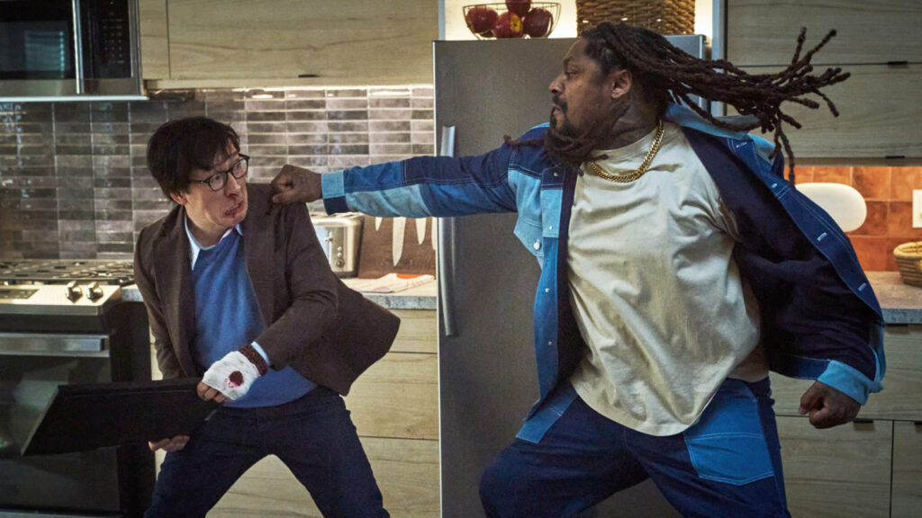 Two men in a kitchen: one with a bandaged hand holding a laptop, dodging a punch from the other man with dreadlocks, wearing a denim jacket.