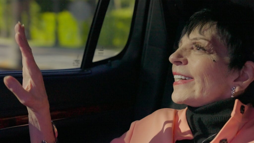 Person with short dark hair and a pink jacket sitting in a car, looking out the window with a smile.