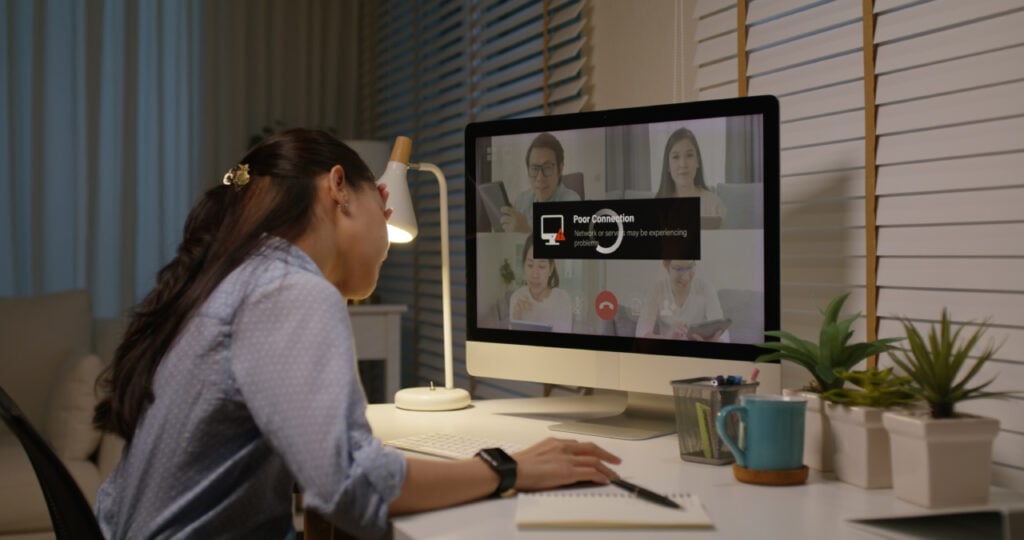 Person using a computer for a video call in a dimly lit room. Screen shows a connection issue notification with other participants visible. Desk has plants, a notebook, and a lamp.