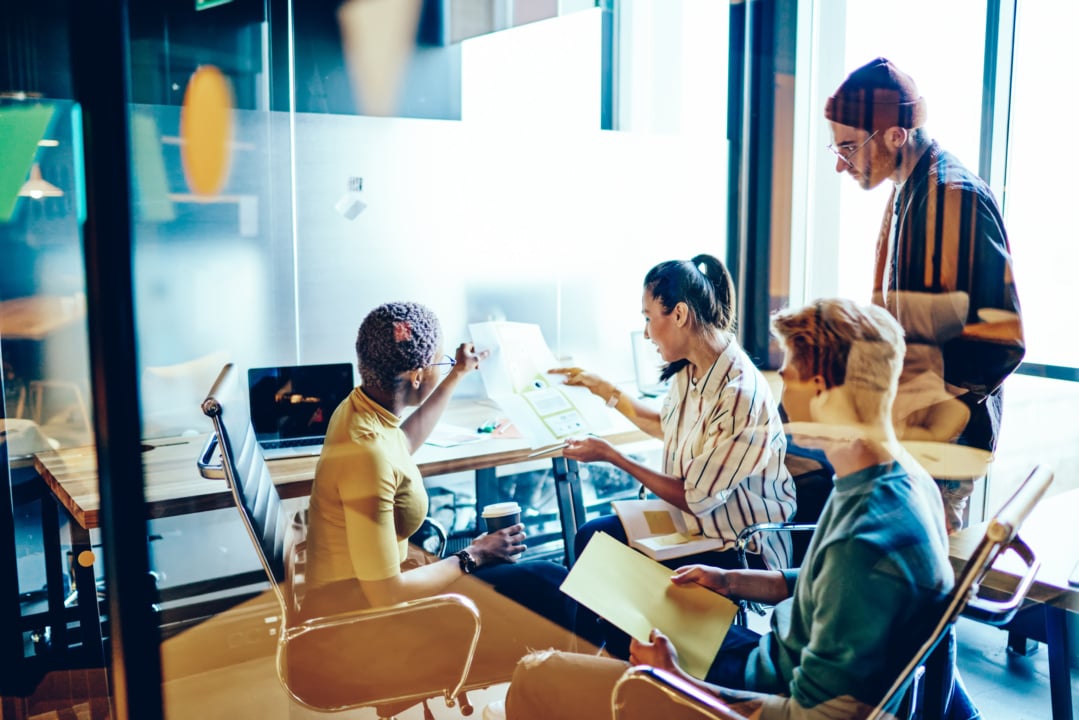 Four people in a modern office setting having a discussion around a table, with laptops open. One person is pointing at a document while the others listen attentively.