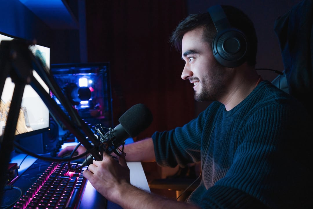 Person wearing headphones, smiling while using a computer and microphone setup.