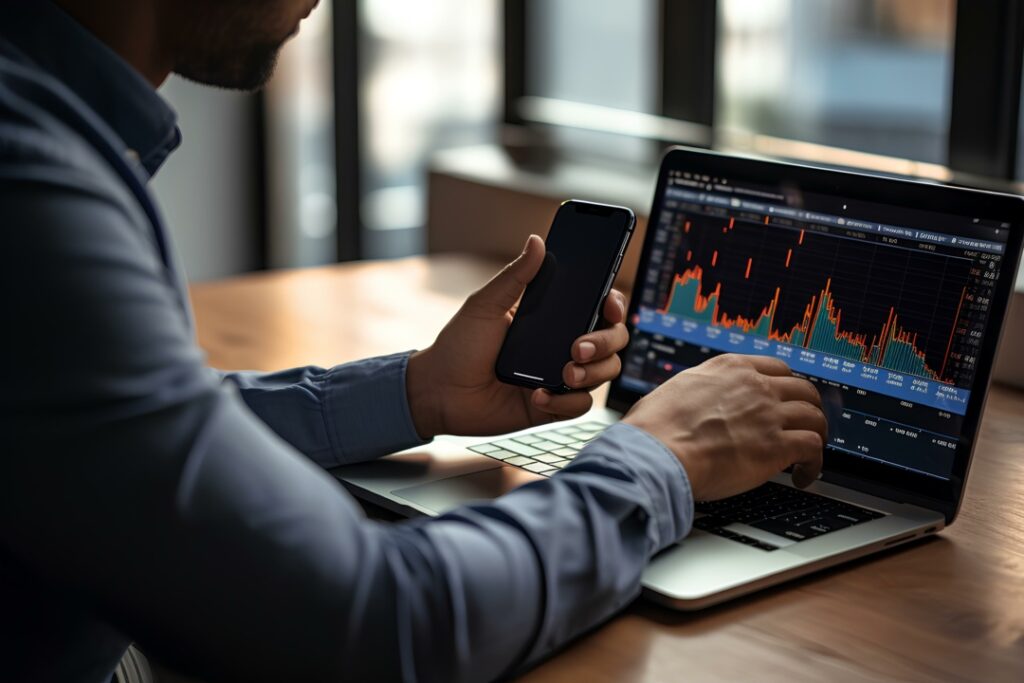 A person analyzing financial data on a laptop, holding a smartphone. The screen displays a graph with rising and falling points.